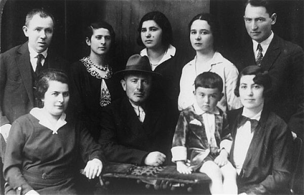 Yitzhaks Familie in Wilna, um 1930. Rechts im Bild sitzt Yitzhak als kleiner Junge auf dem Tisch, neben ihm seine Mutter Rosa, dahinter steht Yitzhaks Vater Eli. Auch die Familie von Yitzhaks Onkel, Zvi Yaakov (der Mann mit Hut in der Mitte des Bildes) ist zu sehen: Yitzhaks Cousinen Rosa (die Zweite von links) und Yona (die Zweite von rechts).